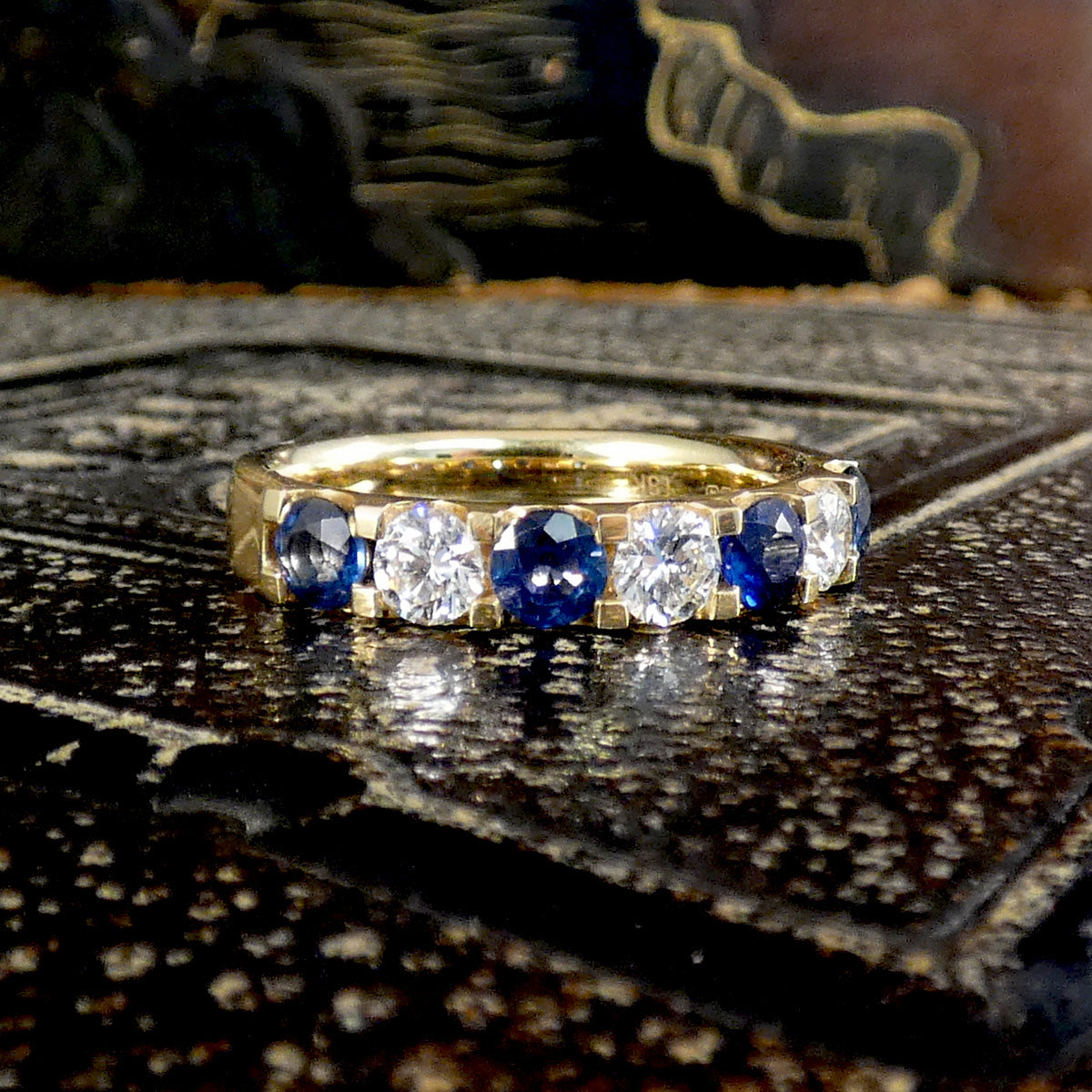 A yellow gold eternity ring featuring alternating round-cut blue sapphires and clear diamonds, resting on a dark, textured metallic surface.