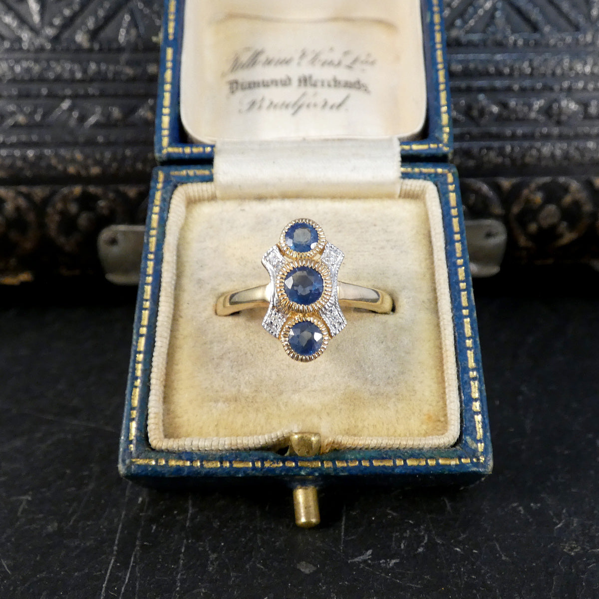 A vintage gold ring with three round blue sapphires and diamond accents, displayed inside an open antique blue velvet jewelry box.