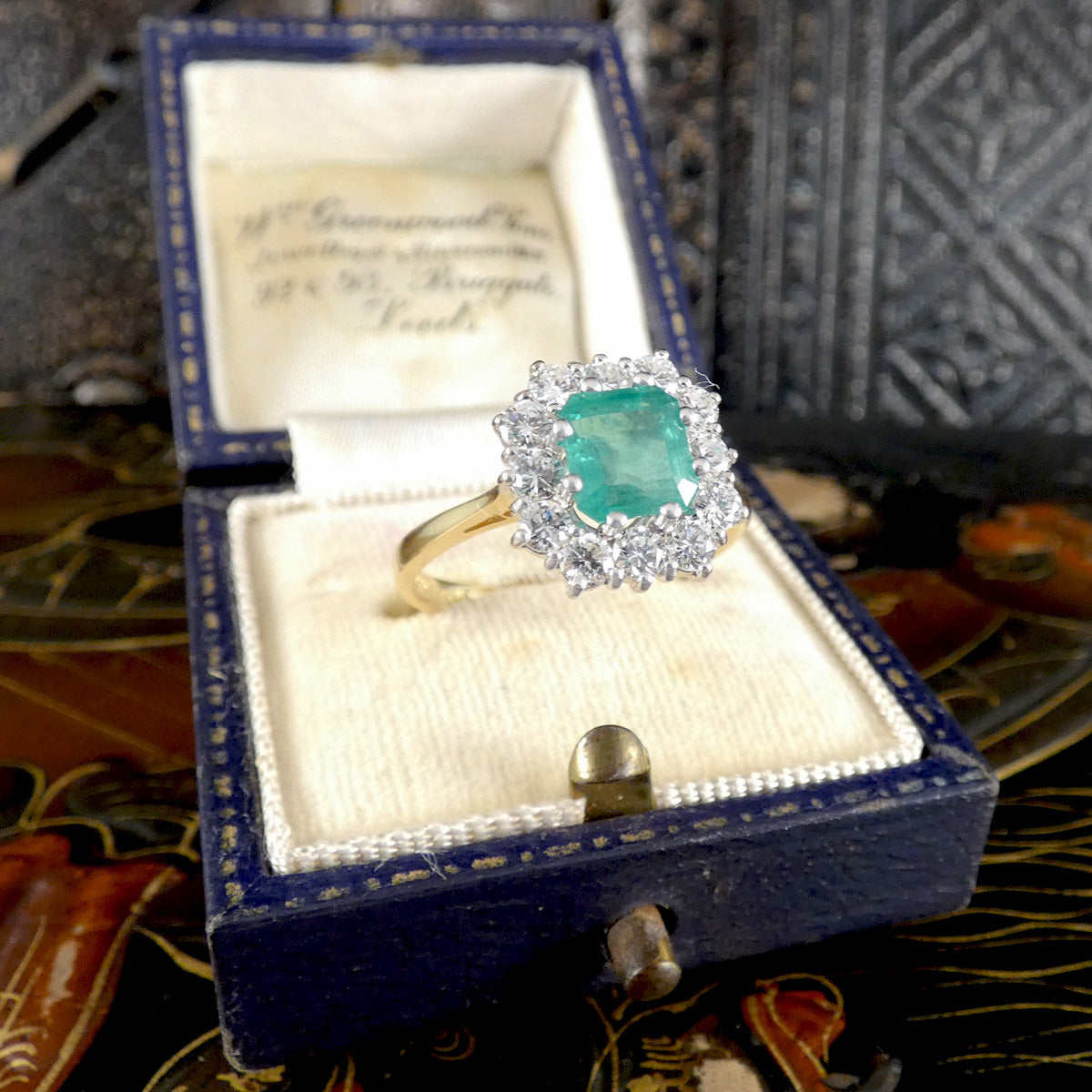 A vintage-style emerald-cut green gemstone ring with a diamond halo and gold band, displayed inside an open dark blue velvet jewelry box.