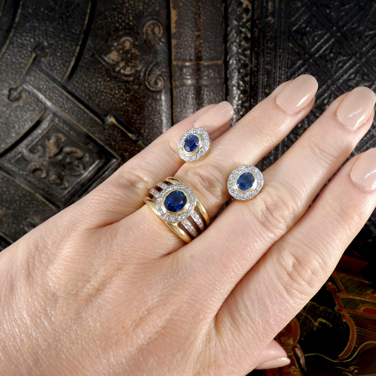 A beautifully bright pair of sapphire and diamond stud earrings in 18ct gold with a bezel setting. Showing them sat on the hand with their matching ring.