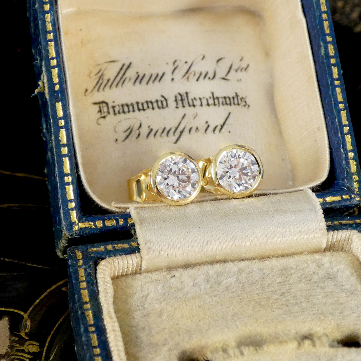 A pair of yellow gold rings with round clear gemstones displayed in an open vintage blue and gold jewelry box with cream velvet lining. The inside lid is printed with 'Fullwood & Co Ltd Diamond Merchants, Bradford.