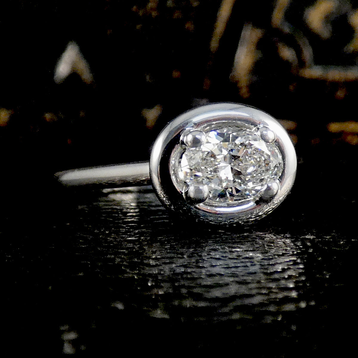 A classic solitaire ring featuring a ovak brilliant-cut diamond set in a polished silver-colored metal bezel, resting on a dark reflective surface.