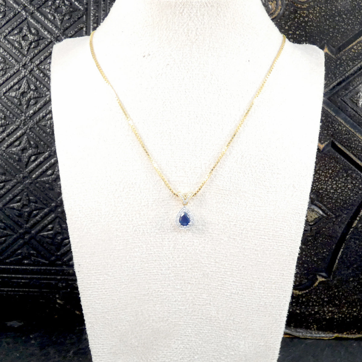 A delicate gold chain necklace with a teardrop-shaped blue gemstone pendant, displayed on a white jewelry bust against a dark textured background.