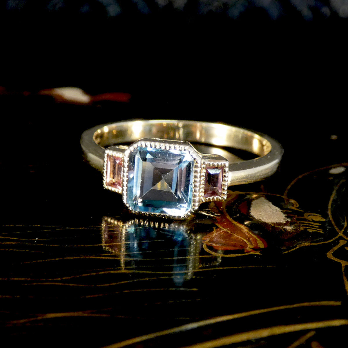 A gold ring featuring a large square-cut light blue gemstone in a beaded halo, flanked by smaller pink and purple accent stones, resting on a dark reflective surface.