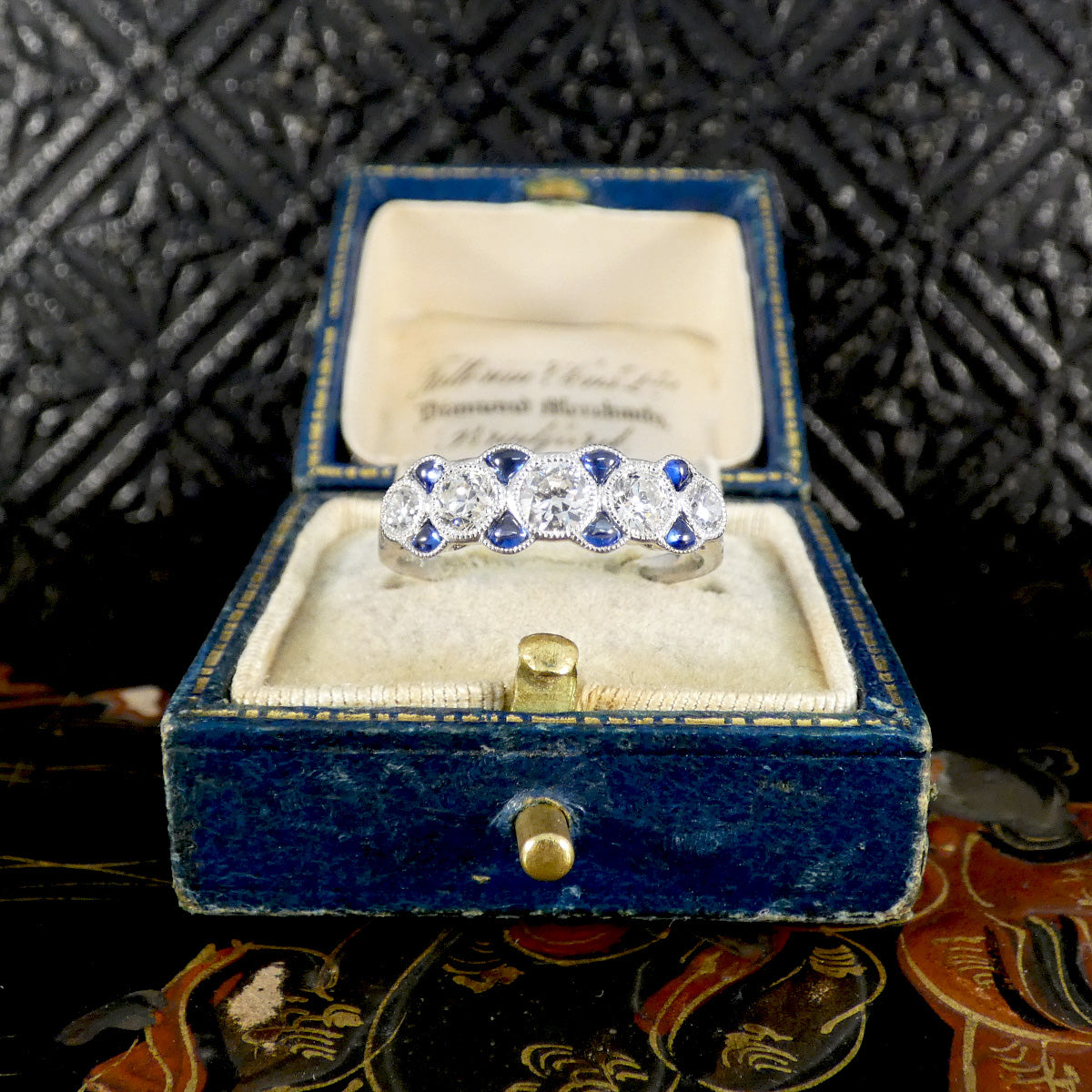 Art Deco-inspired platinum ring with five old-cut diamonds and vivid blue sapphire accents, elegantly presented in a vintage blue velvet ring box.