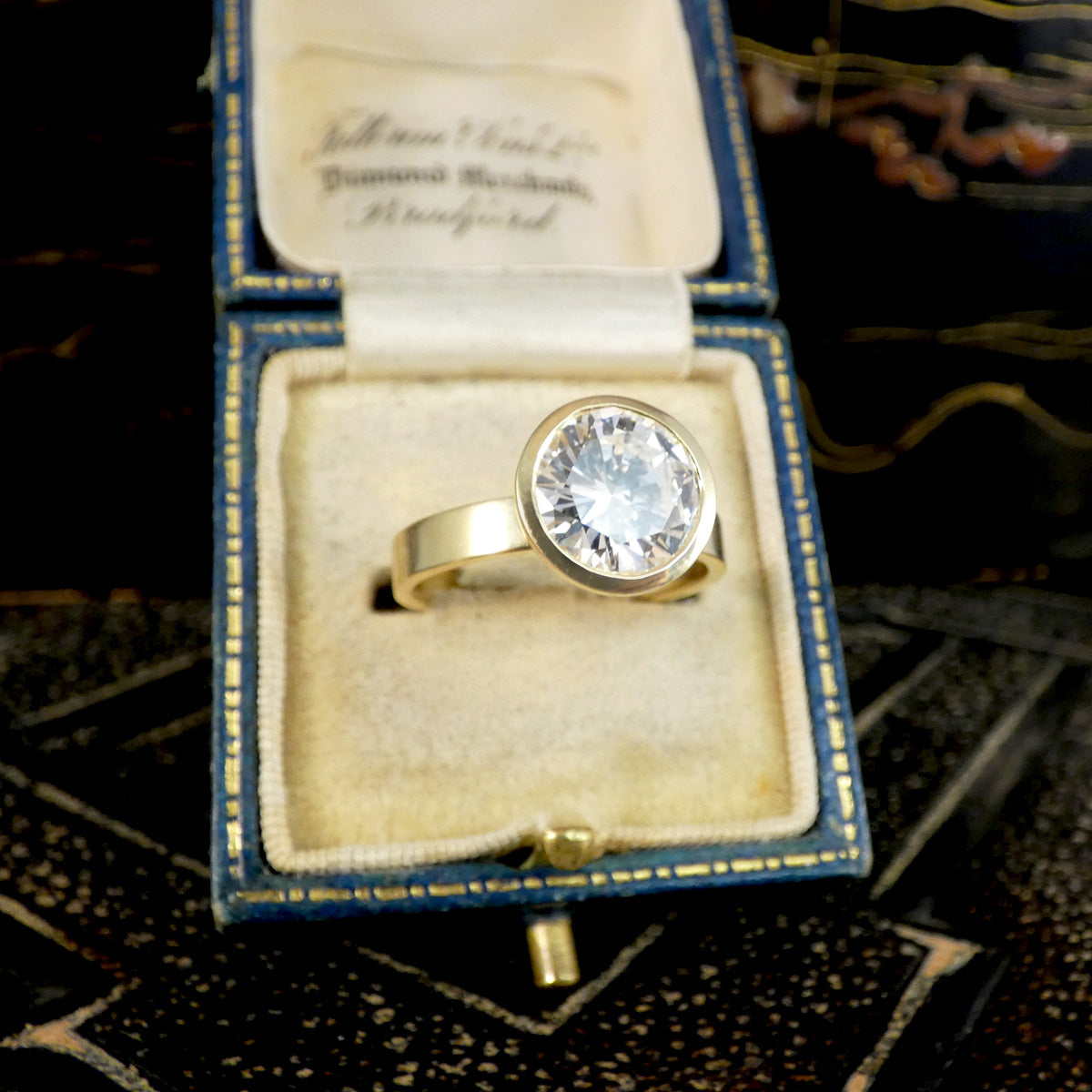 A diamond solitaire ring in a rub over bezel setting in 18ct yellow gold shown in jewellery box