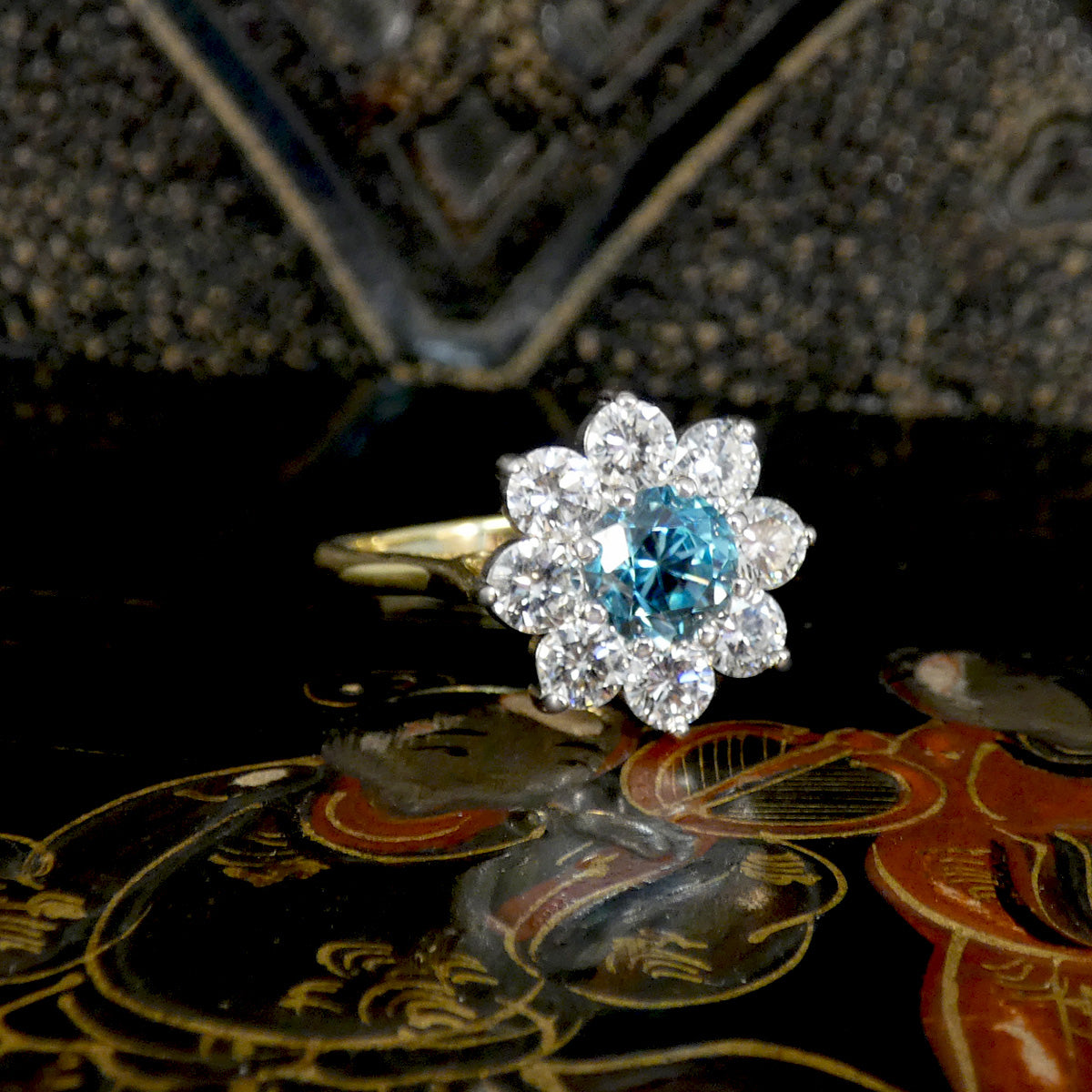 A floral cluster ring featuring a central round blue gemstone surrounded by clear stones, set on a gold band and photographed against a dark decorative background.
