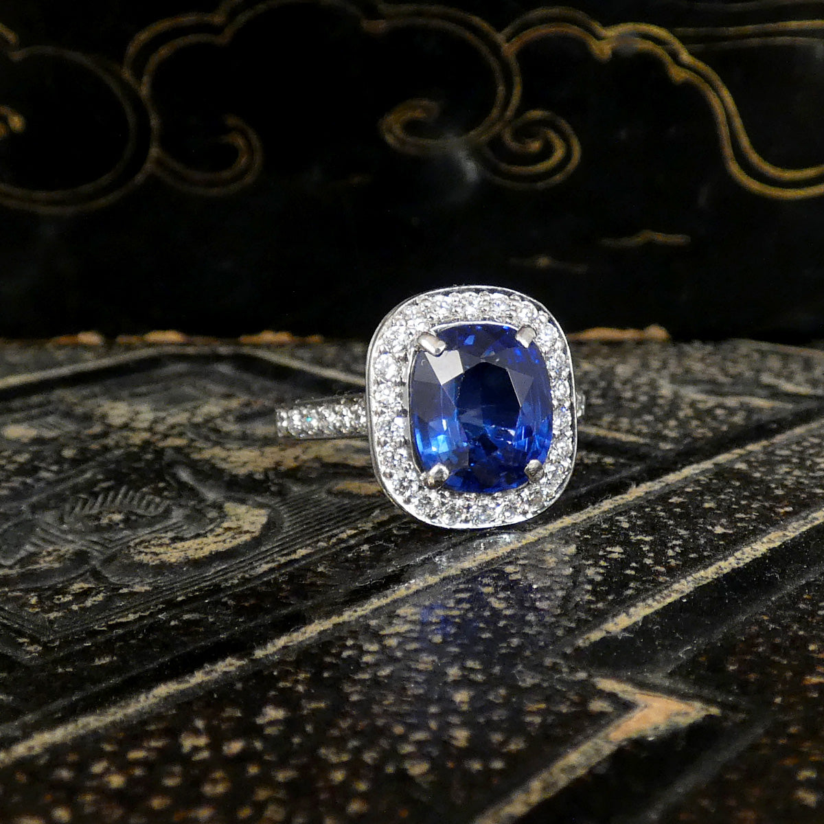 A silver-toned halo ring featuring a large oval-cut deep blue sapphire center stone surrounded by small clear stones, displayed on a dark patterned surface.