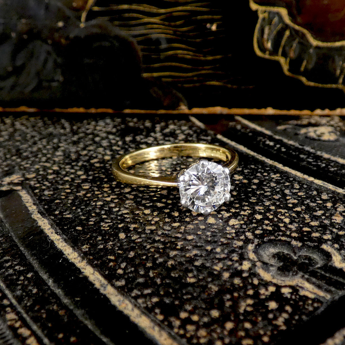 A yellow gold ring with a single round-cut clear gemstone, resting on a textured black and gold surface.