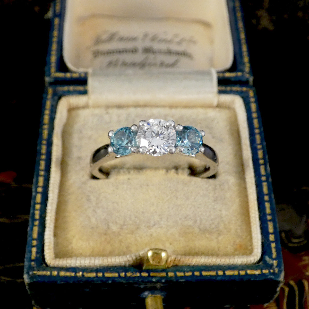A three-stone diamond engagement ring with a round center stone and two round blue side stones, displayed in an open vintage blue and gold ring box with a cream velvet insert.