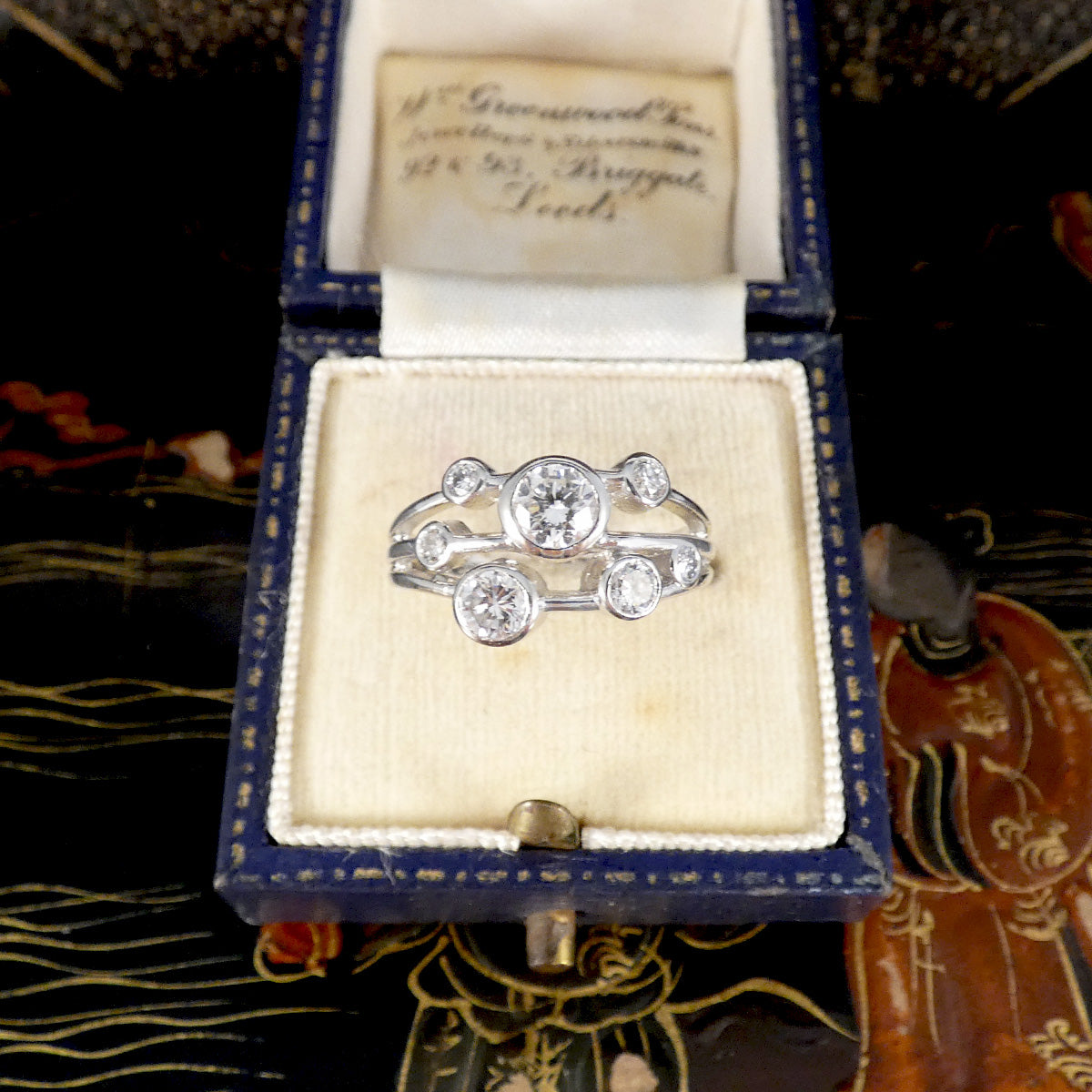 A vintage silver ring with a central round diamond and smaller surrounding diamonds, displayed inside an open antique blue velvet ring box with a cream insert.