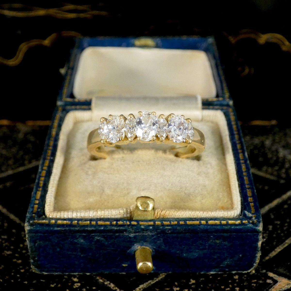 A yellow gold five-stone diamond ring displayed inside an open vintage blue velvet ring box with a cream satin lining, set against a dark patterned background.