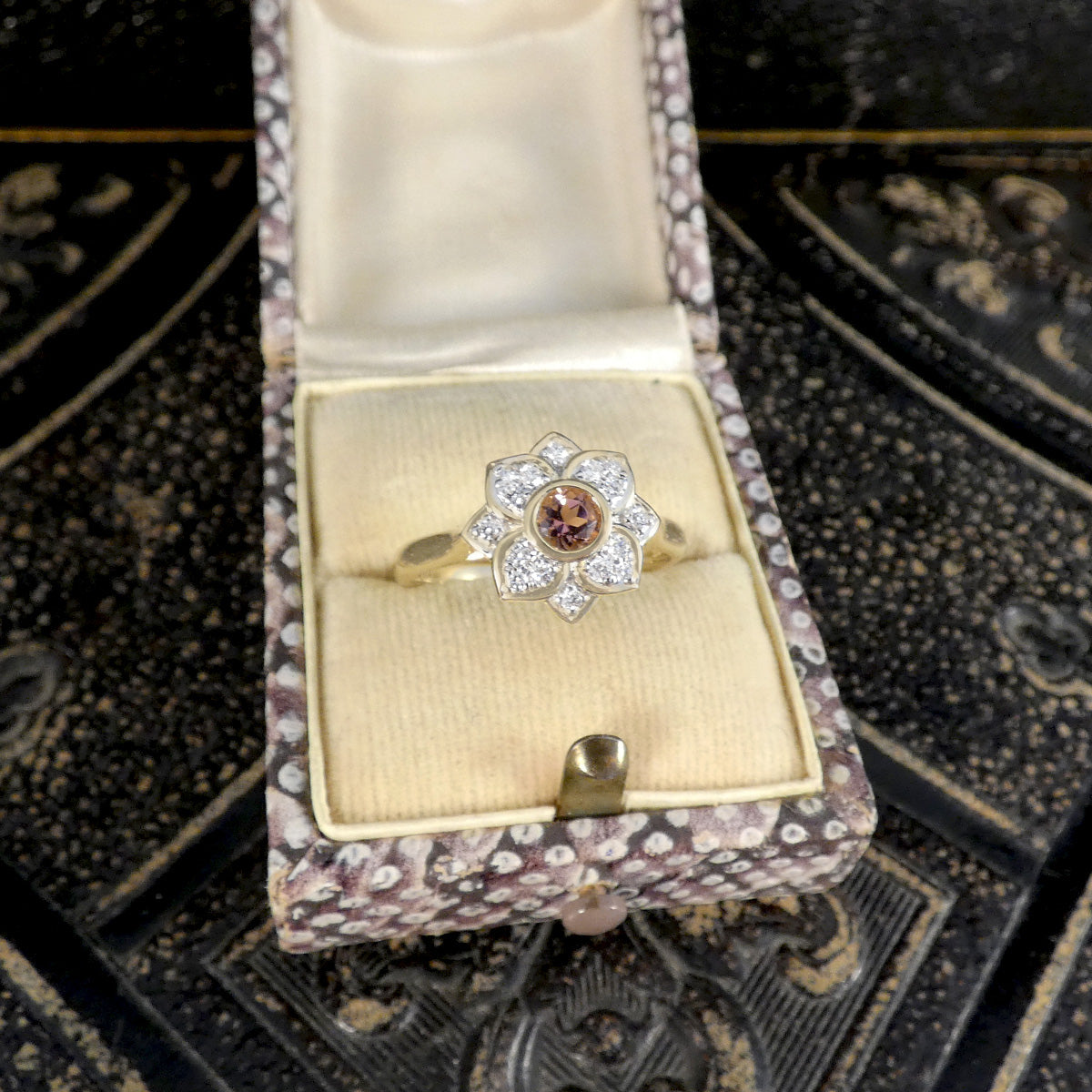Gold ring with a diamond and pink gemstone in an open jewelry box on a decorative surface.