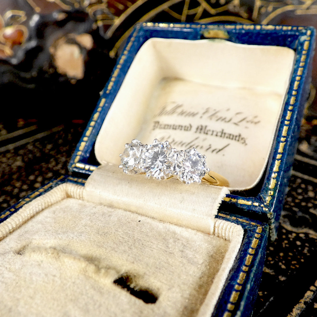 A three-stone diamond ring with a yellow gold band displayed inside an open vintage blue velvet ring box with gold trim and cream lining.