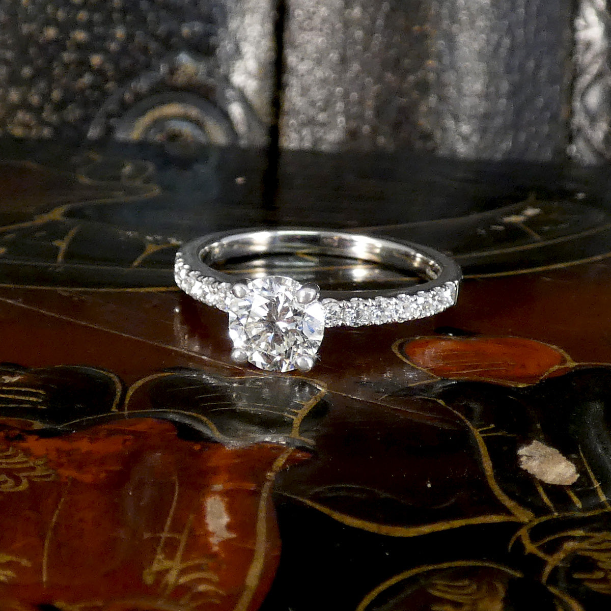 A silver-toned diamond engagement ring with a round-cut center stone and pavé-set side stones, resting on a glossy dark brown wooden surface with a textured metallic background.