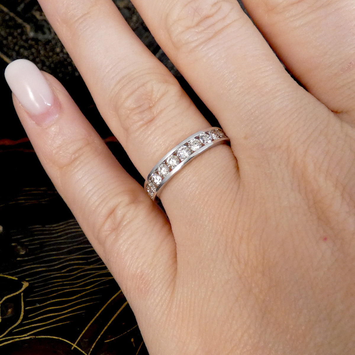 A close-up of a hand wearing a white gold eternity ring set with small diamonds. The hand has light pink manicured nails, and the background is a dark textured surface with gold lines.