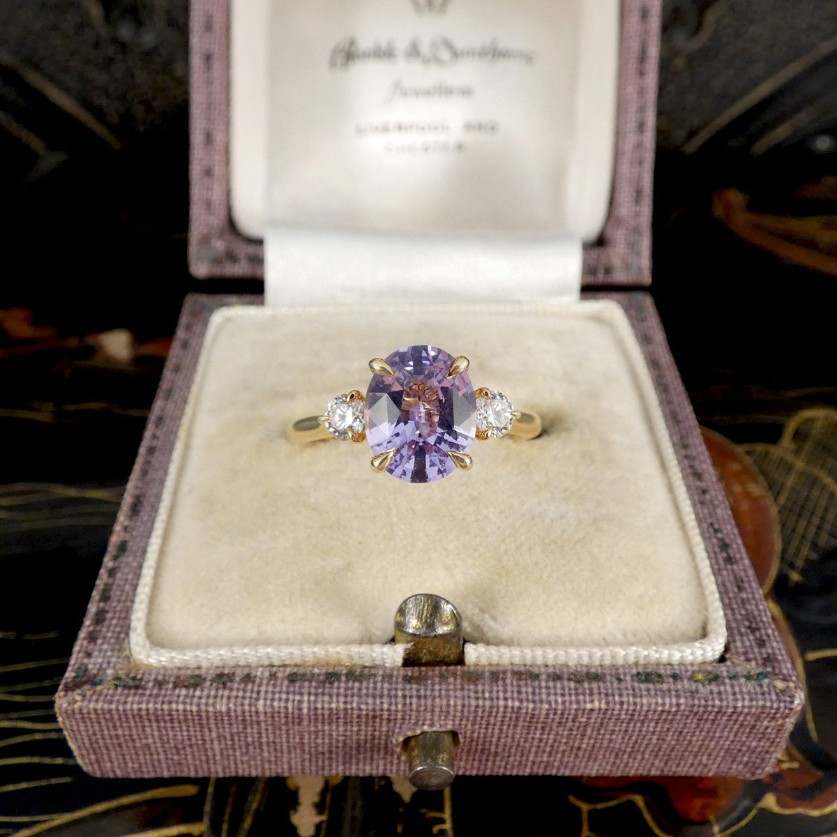 A yellow gold ring with a central oval purple sapphire gemstone and two round diamond accent stones, displayed in an open vintage lavender velvet ring box.