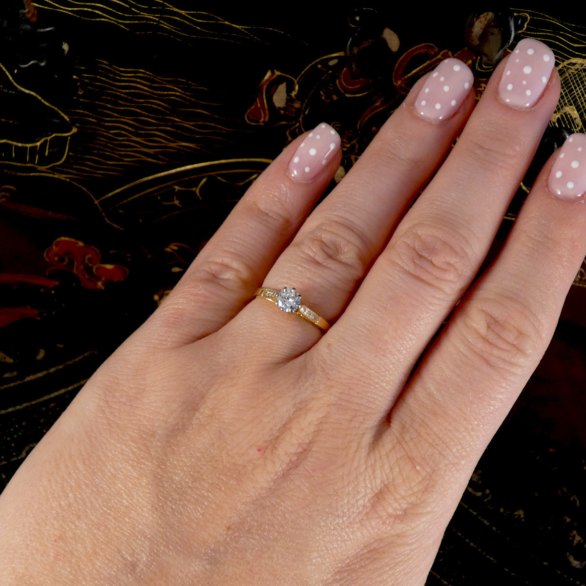 A close-up of a hand wearing a yellow gold ring with a round-cut clear gemstone, set against a dark patterned background.