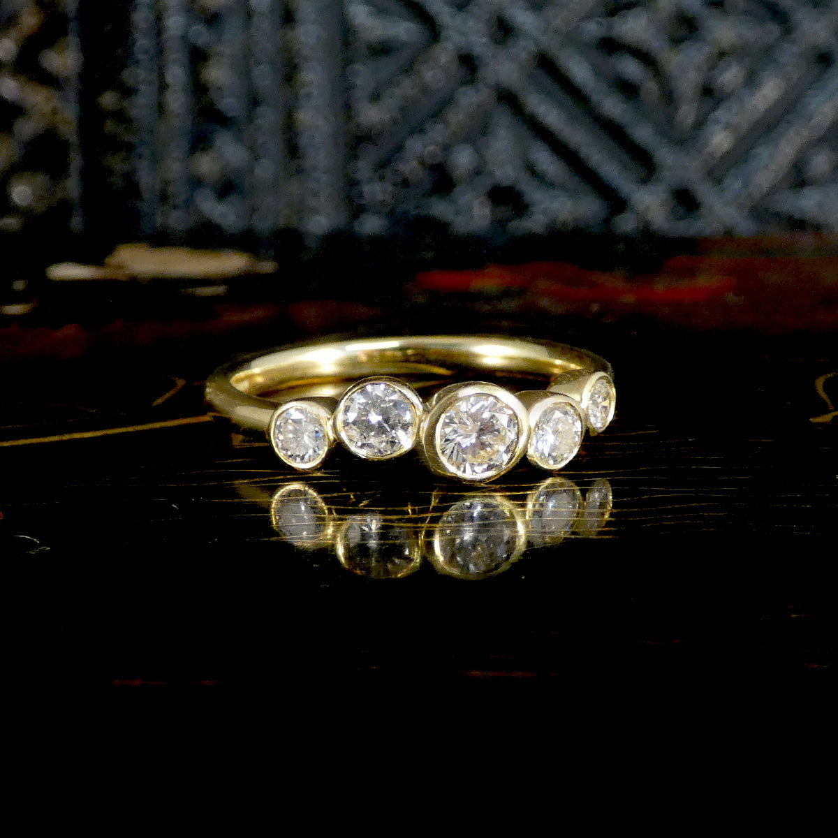 A yellow gold ring featuring five round-cut clear gemstones in bezel settings, displayed on a dark reflective surface with a blurred dark background.