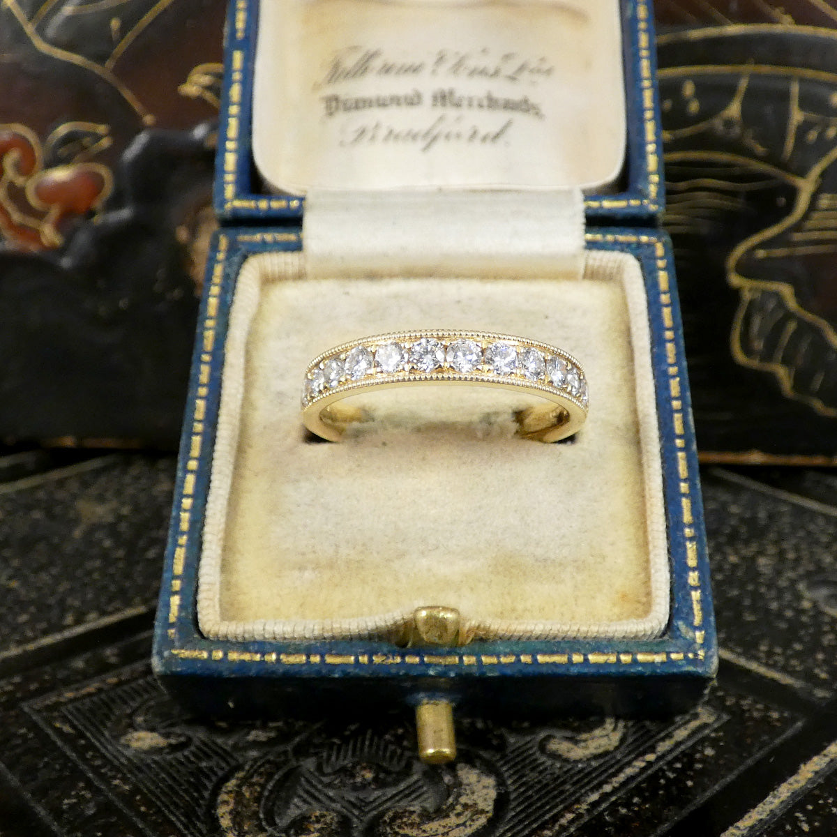 A yellow gold eternity ring with a row of clear diamonds set in an open vintage blue velvet ring box. The box has gold trim and a cream-colored interior with cursive writing on the inner lid.