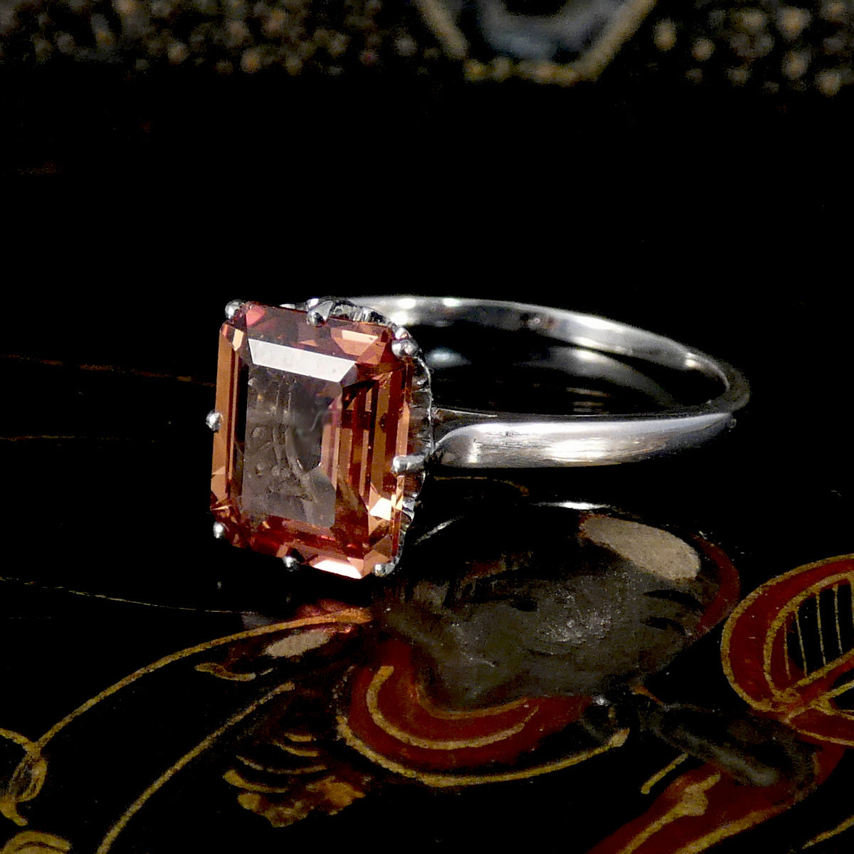 A silver ring featuring a rectangular peach-colored gemstone with a faceted, stepped cut, set against a dark reflective background.