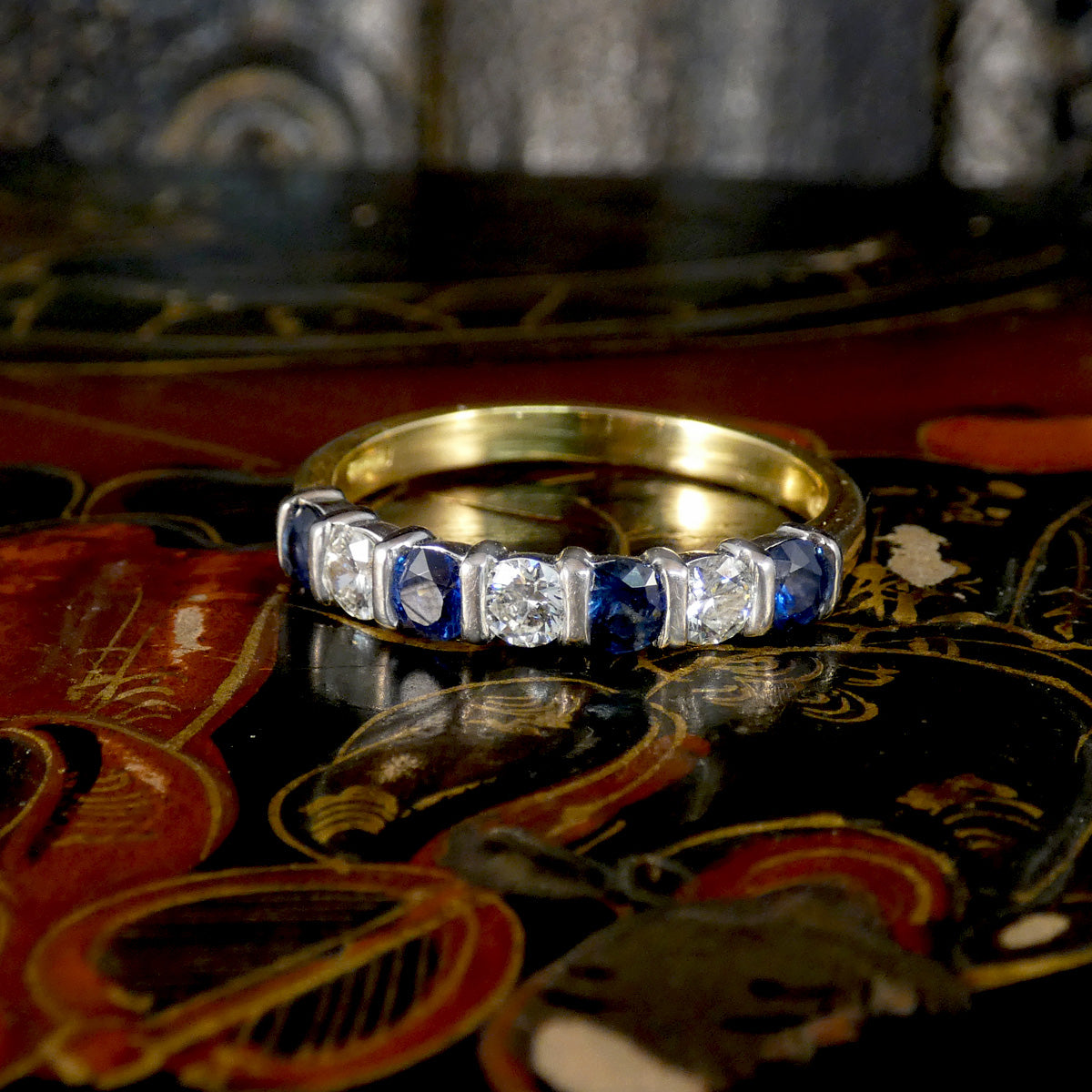 A close-up of a gold band ring set with alternating round-cut blue sapphires and clear diamonds, resting on a dark, ornate surface.