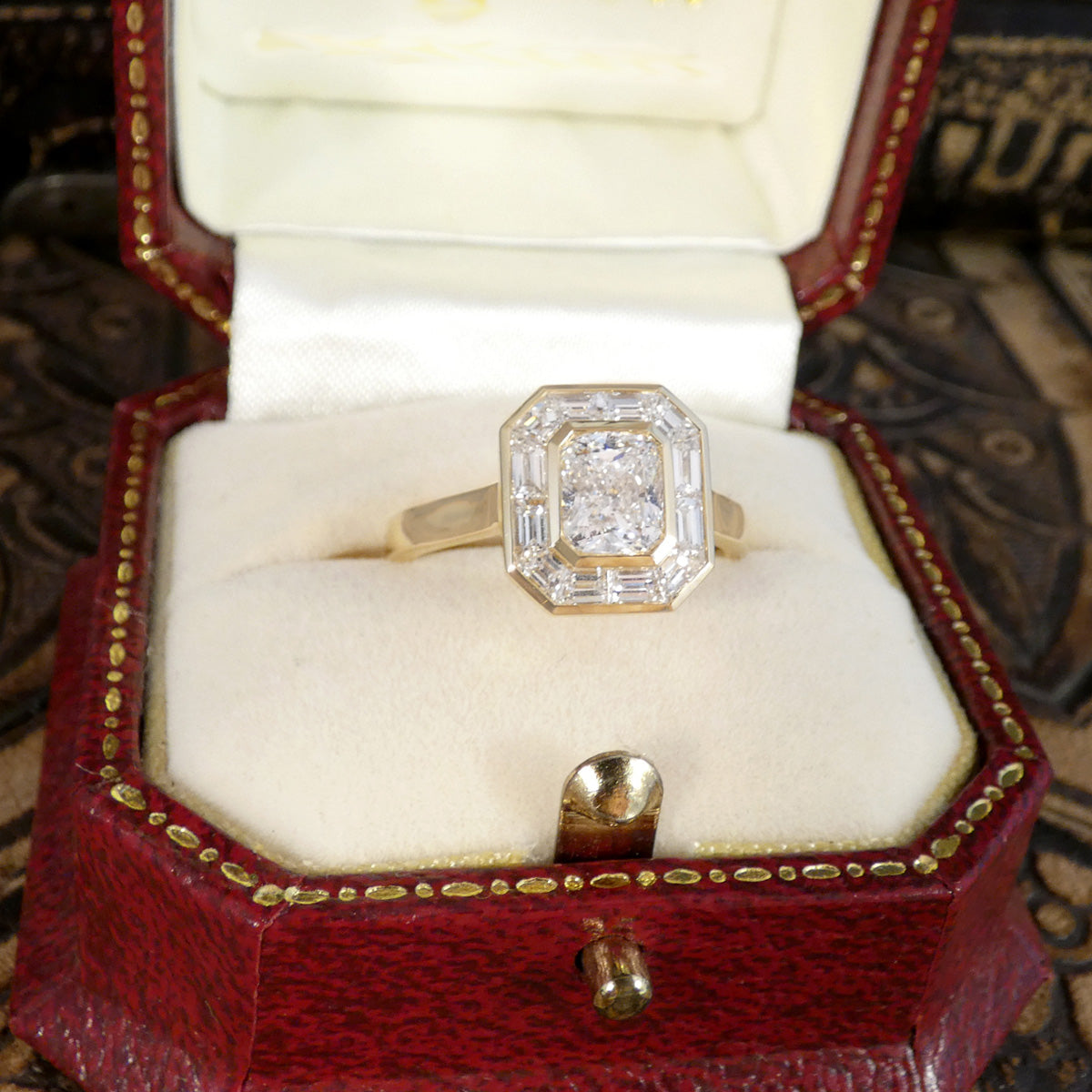A yellow gold ring with a baguette-cut diamond and a smaller round center stone, displayed in an open vintage red ring box with a cream velvet insert.