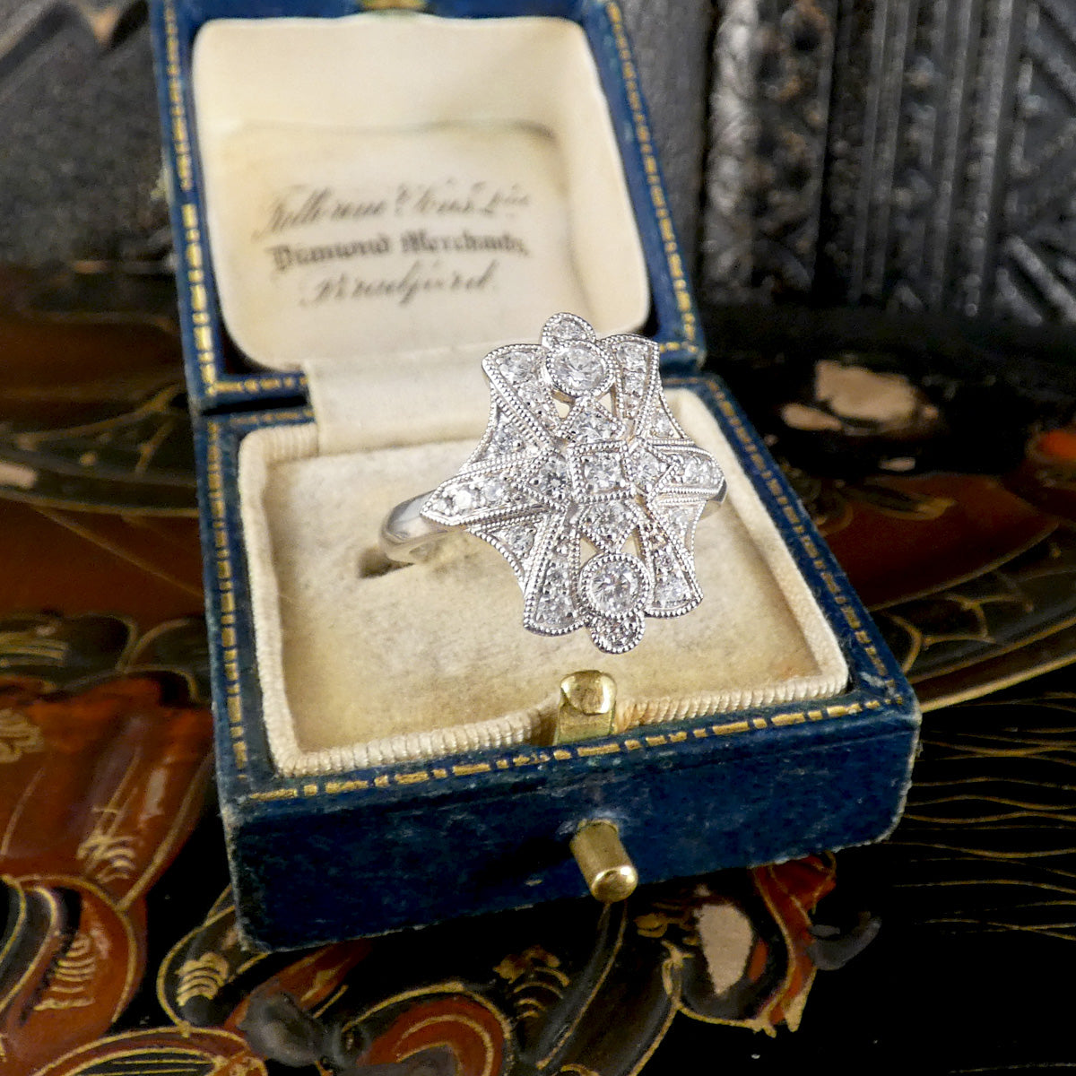 A platinum filigree ring with clear diamond stones, displayed inside an open vintage blue velvet ring box with a cream velvet interior.