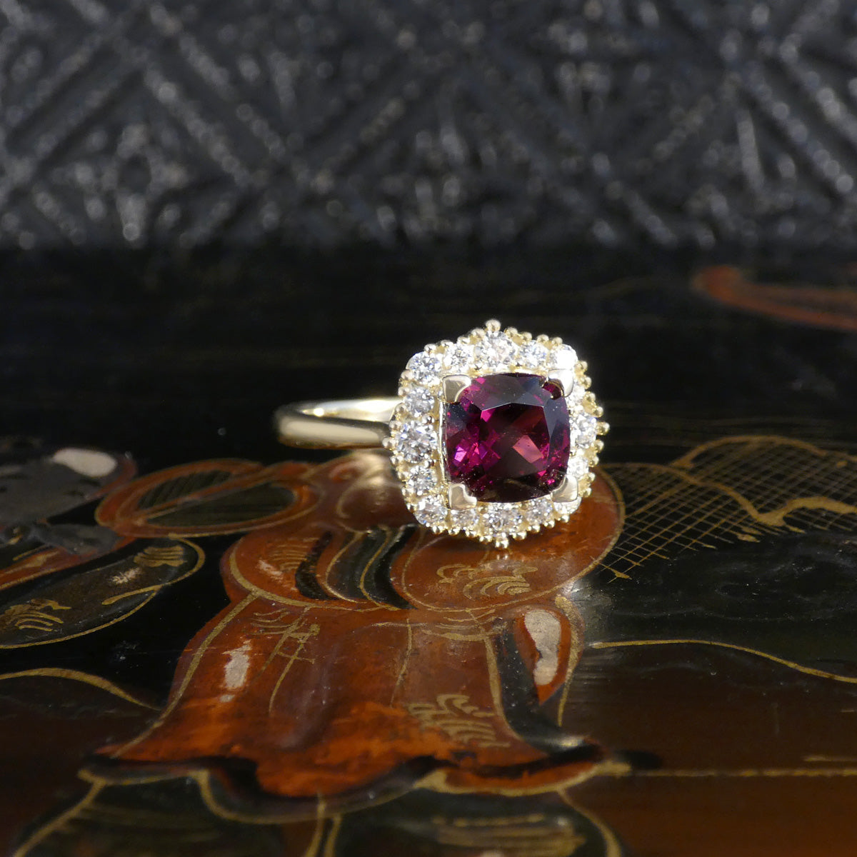 A yellow gold ring featuring a cushion-cut pinkish-red center stone surrounded by a halo of small clear stones, displayed on a dark, patterned surface.