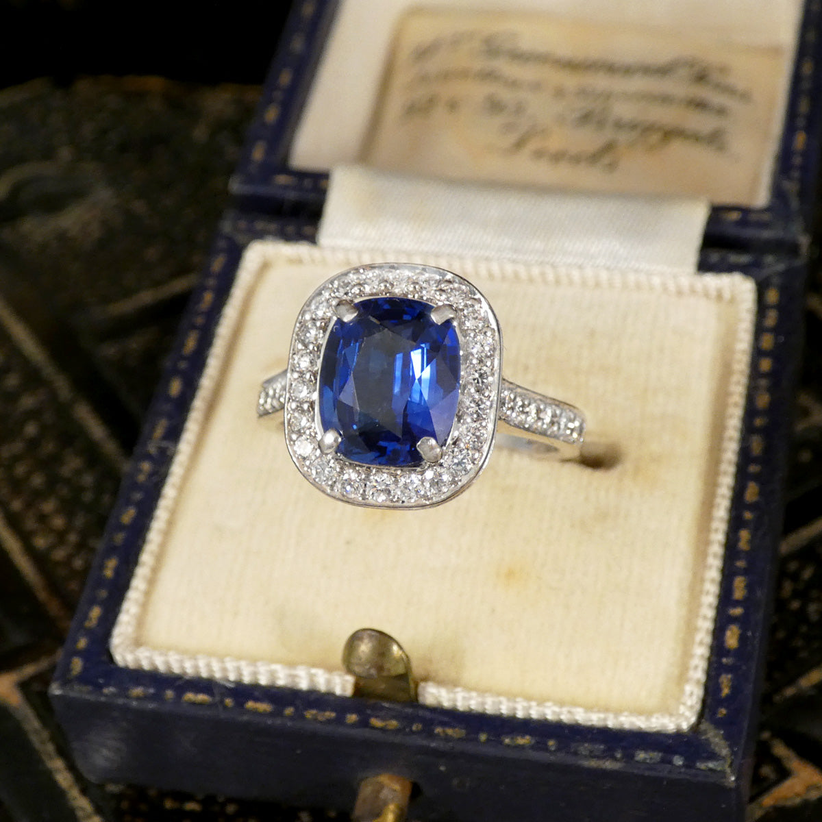 A vintage-style ring with a rectangular blue sapphire and a halo of small clear stones, displayed in an open dark blue jewelry box with a cream velvet insert.