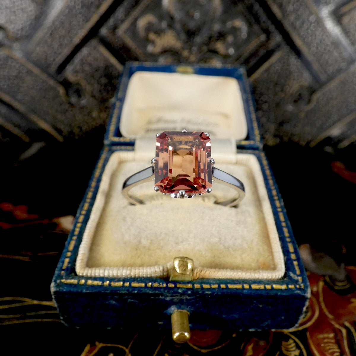 An emerald-cut orange sapphire ring set in a platinum band, displayed inside an open blue velvet ring box with a cream lining.