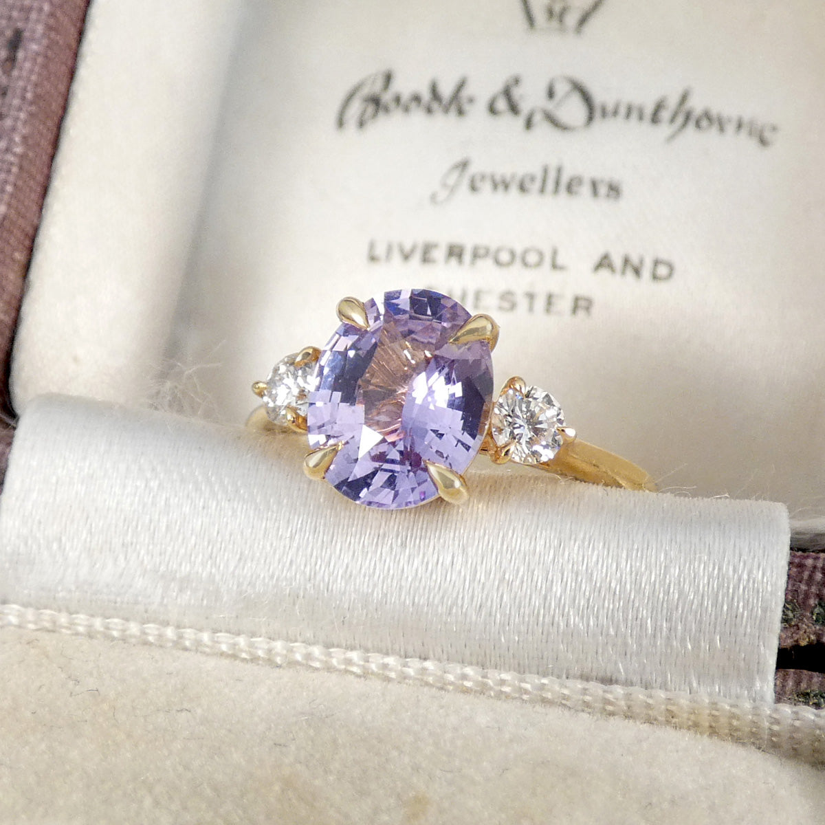 A yellow gold ring with an oval light purple center stone and two small clear accent stones, displayed inside a cream-colored ring box with the brand name 'Boobie & Dunthorne Jewellers' visible on the insert.