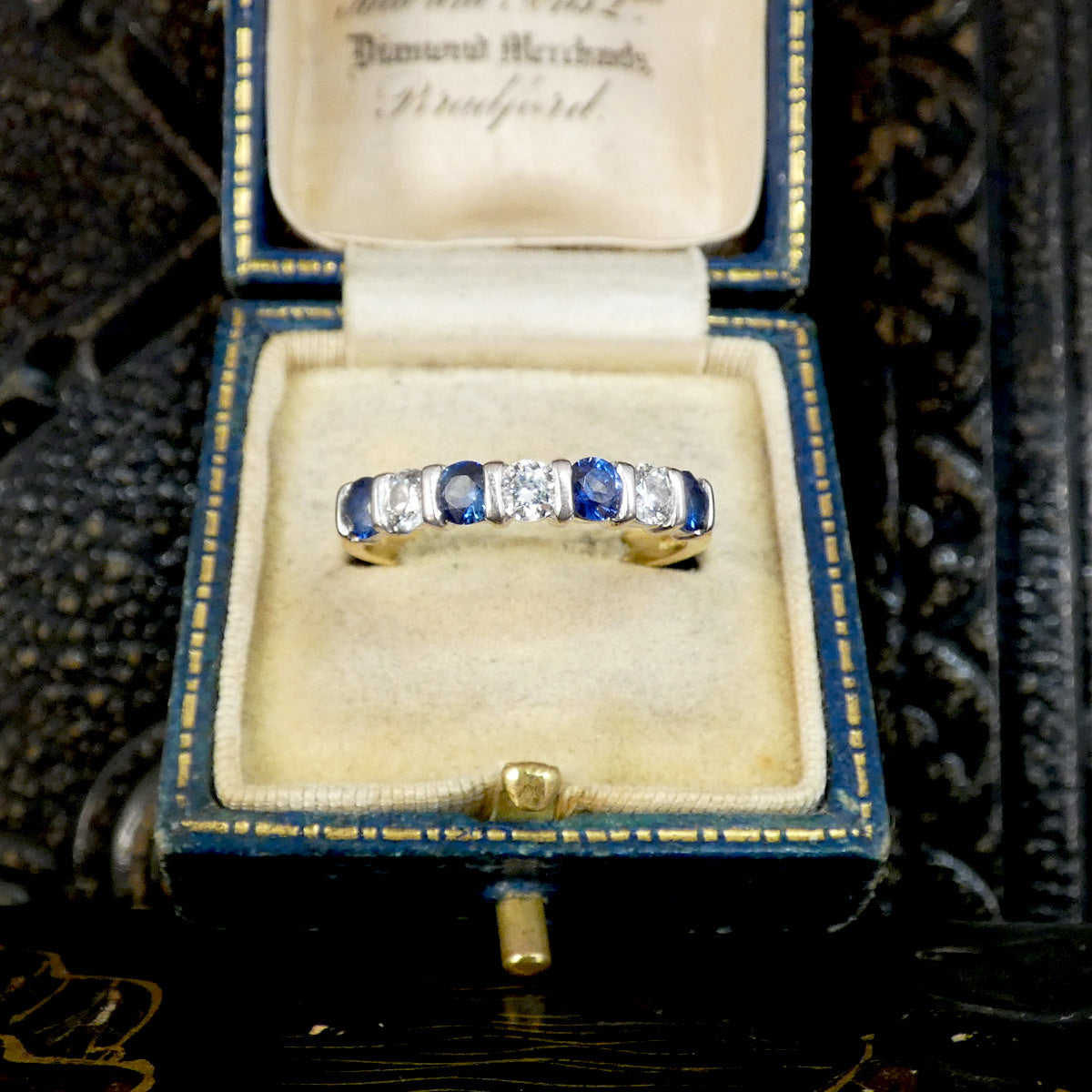 A vintage yellow gold eternity ring with alternating blue sapphires and clear round diamonds, presented in an antique blue velvet ring box.