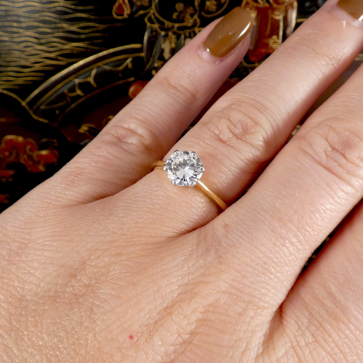 A close-up of a hand wearing a yellow gold solitaire ring with a round-cut clear gemstone. The hand has brown manicured nails, and the background is a dark, ornate patterned surface.