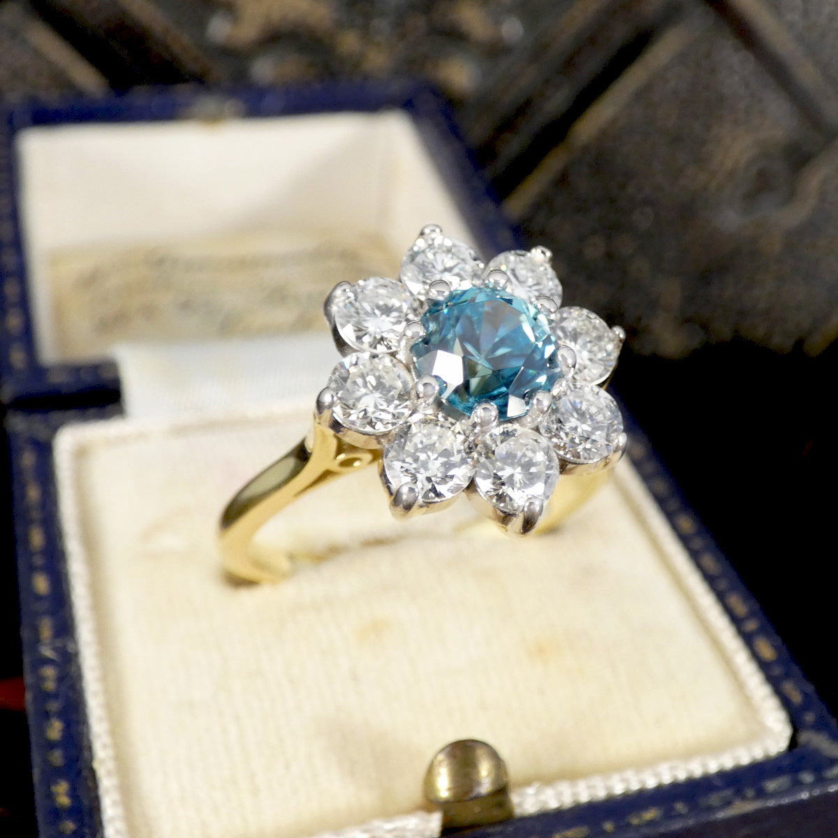 A vintage-style flower cluster ring with a central light blue gemstone surrounded by clear stones, set on a yellow gold band and displayed inside an open blue jewelry box.