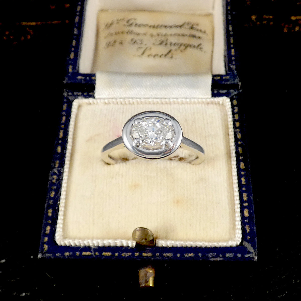 A vintage silver engagement ring with a round diamond solitaire, displayed inside an antique blue and cream jewelry box with an inscription on the inner lid.