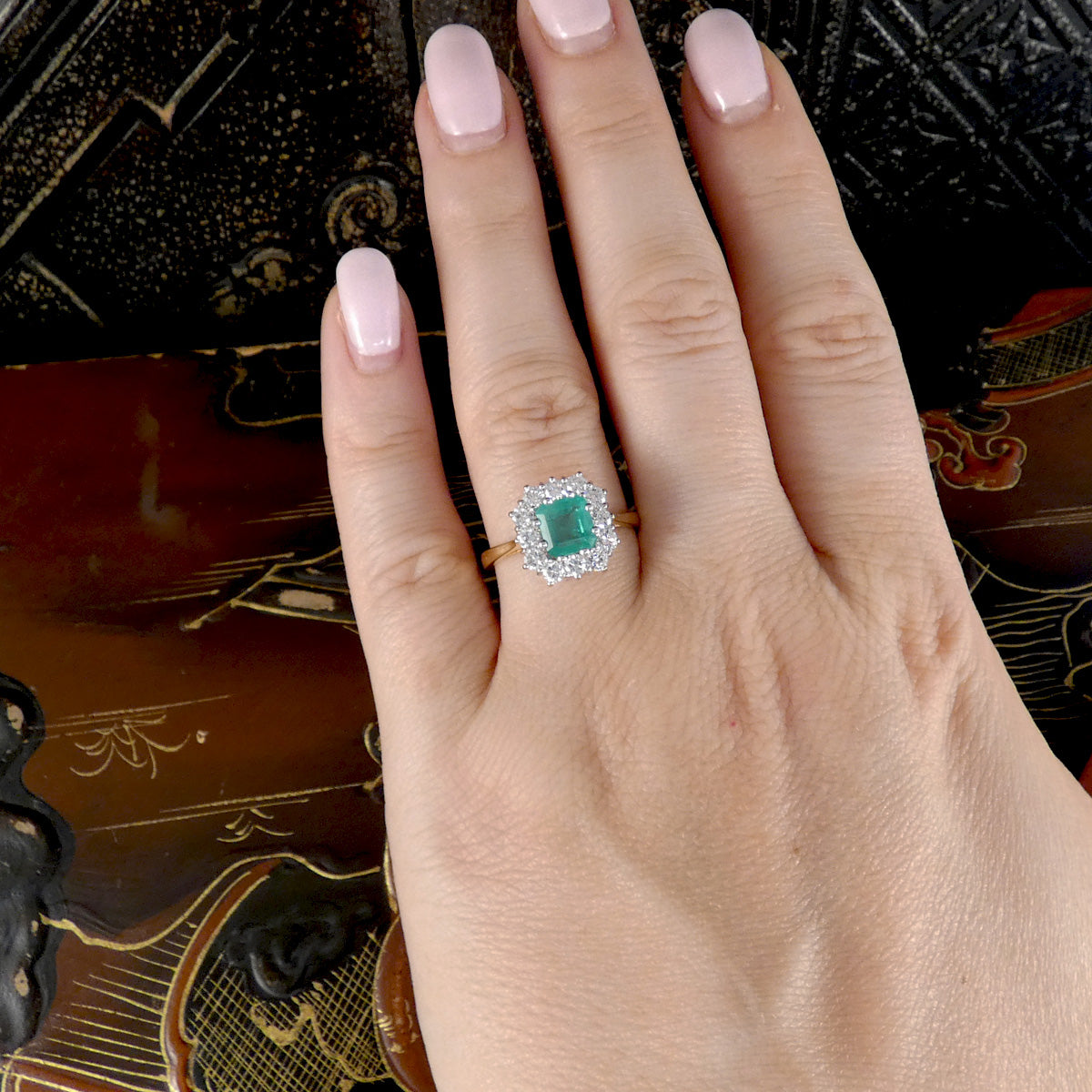 A close-up of a hand wearing a ring with a square-cut green center stone surrounded by a halo of small clear stones on a gold band, set against a dark patterned background.