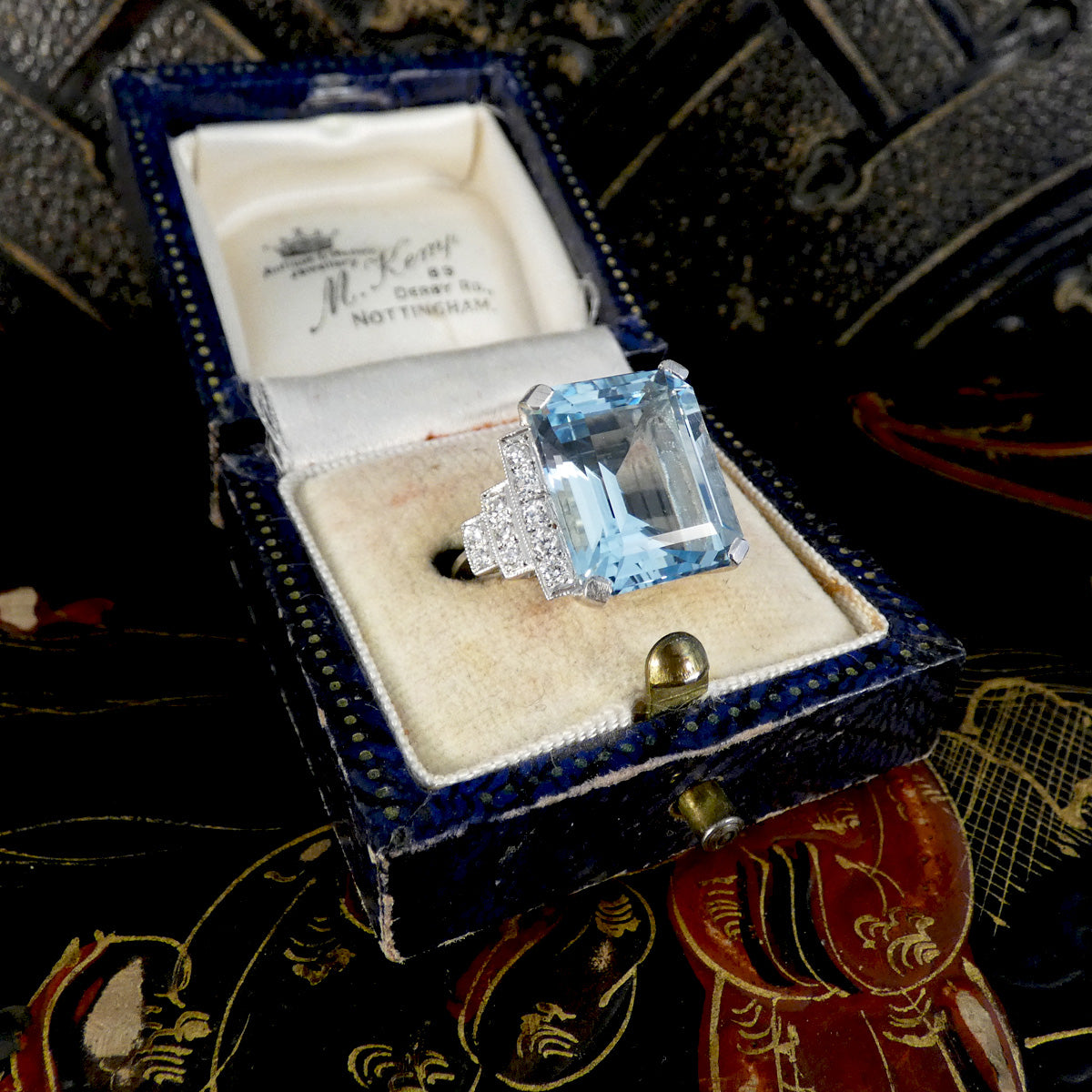 A vintage ring box opened to display a ring with a large light blue emerald-cut gemstone and diamond accents, set against a dark patterned background.