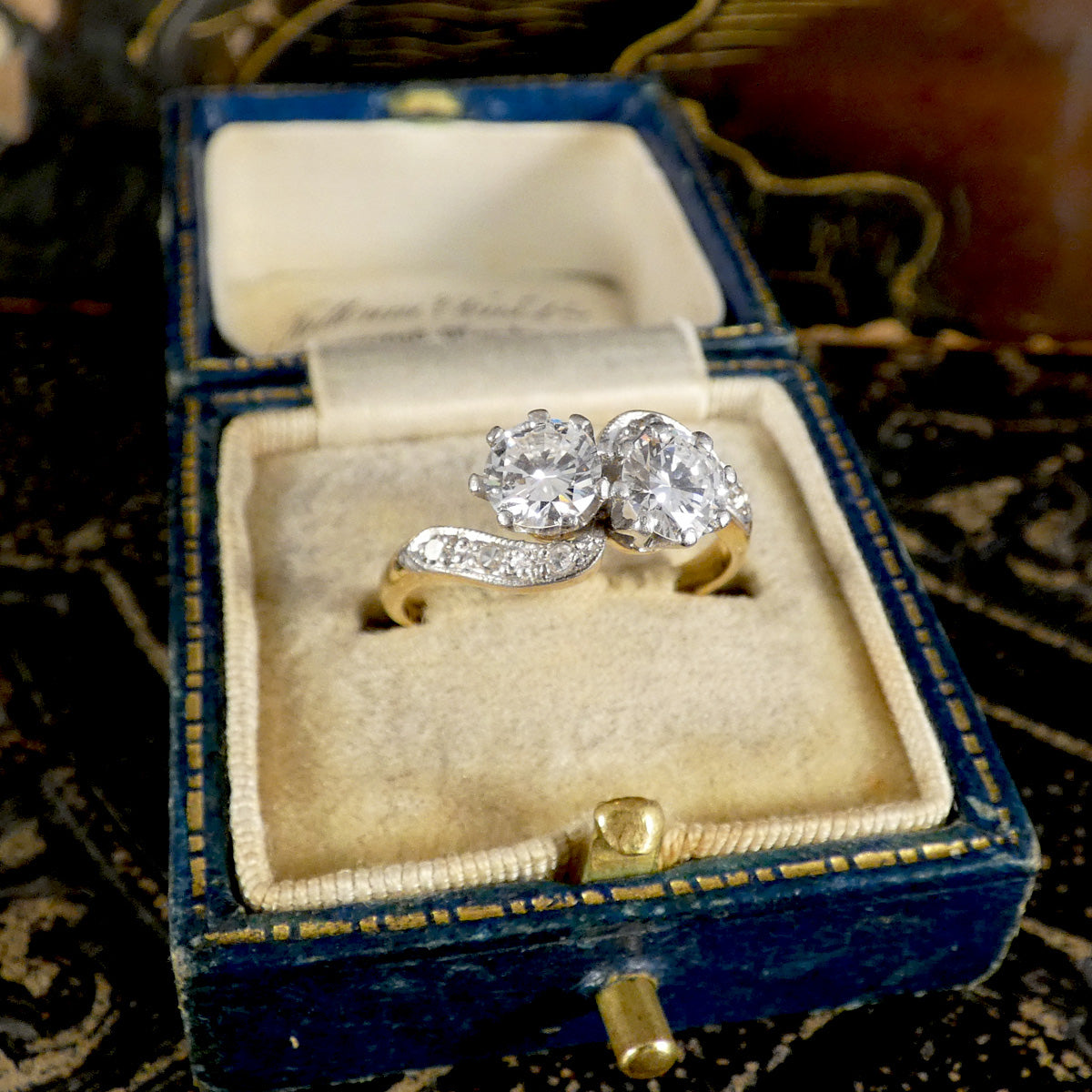 A silver ring with two round-cut clear gemstones and a curved pavé band, displayed inside an open vintage blue velvet jewelry box with a cream insert.
