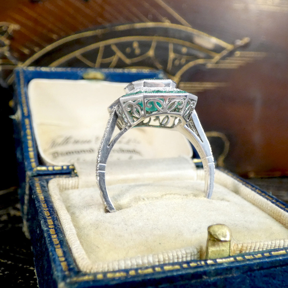Striking Art Deco-style ring featuring a central emerald-cut emerald, surrounded by a double halo of calibre-cut emeralds and pavé-set diamonds, all set in platinum with a full detailed gallery setting.