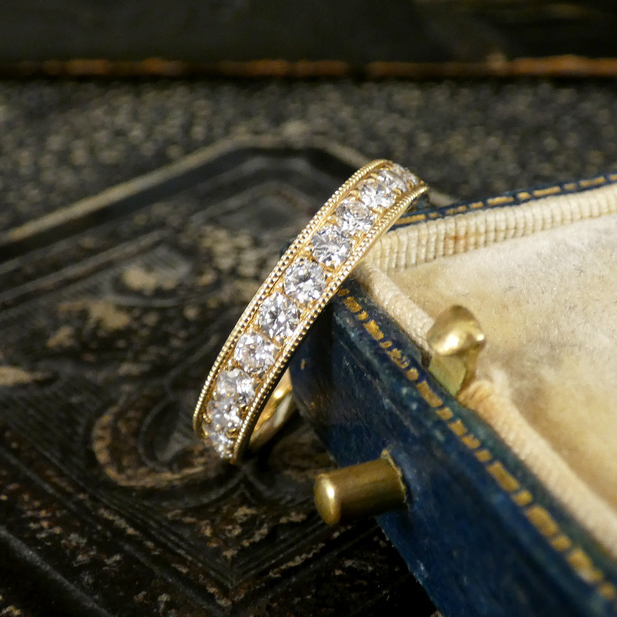 A yellow gold eternity ring set with clear diamonds, resting on an antique blue jewelry box.