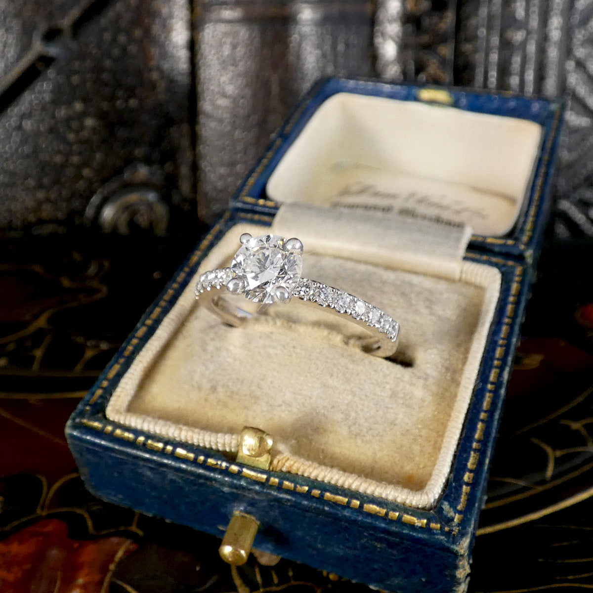 A white gold diamond engagement ring with a round center stone and diamond set band, displayed inside an open vintage blue velvet ring box with gold trim.