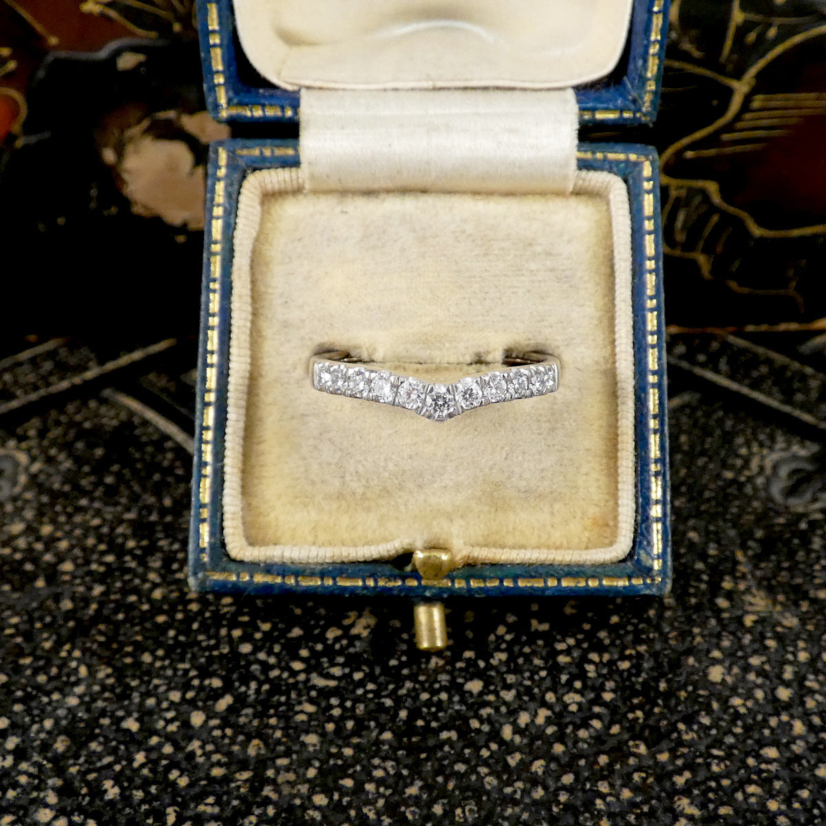 A silver V-shaped ring set with small clear stones, displayed inside an open vintage blue jewelry box with a cream velvet lining.