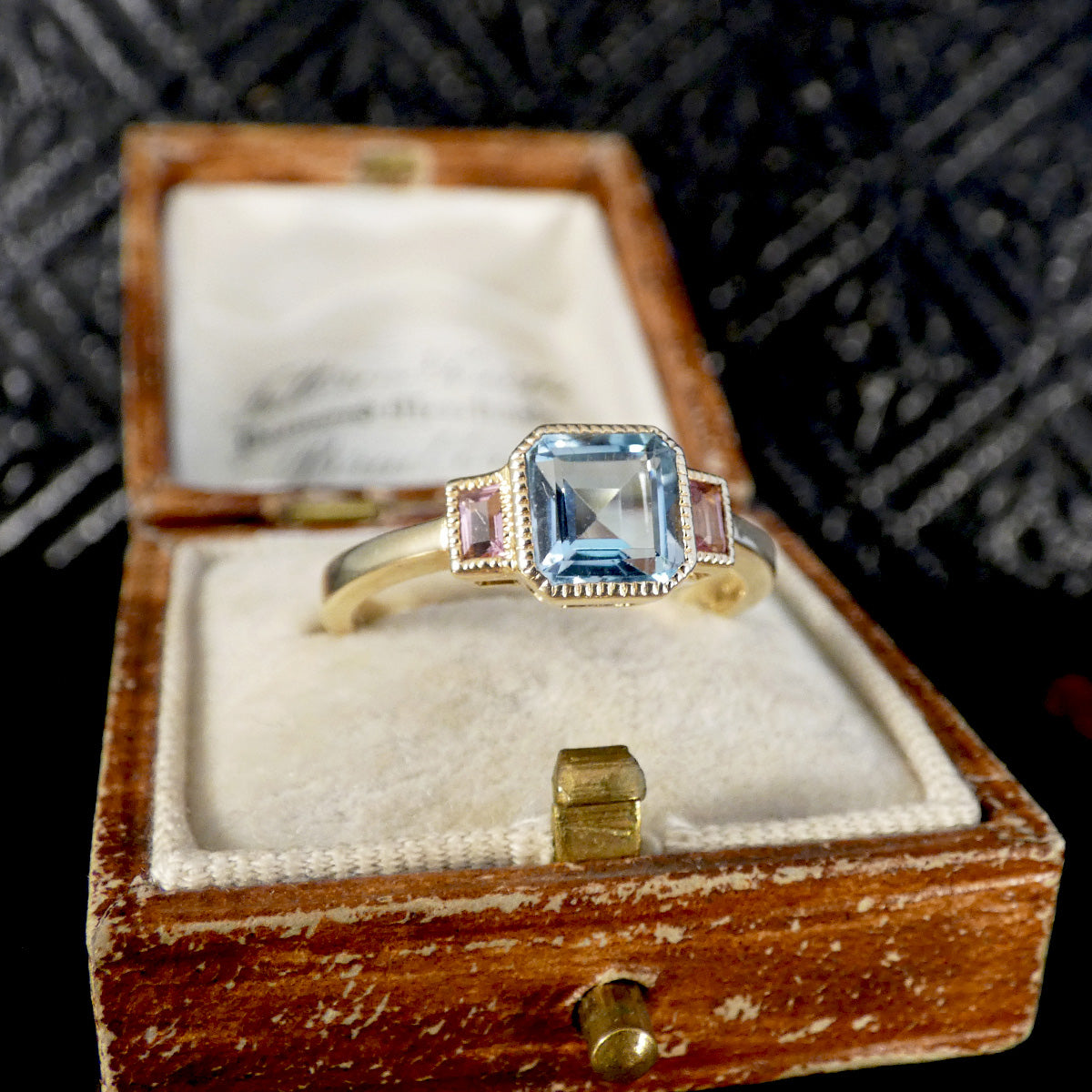 A vintage-style ring with a central light blue cushion-cut gemstone and pink side stones, displayed in an open wooden jewelry box with a cream velvet insert.