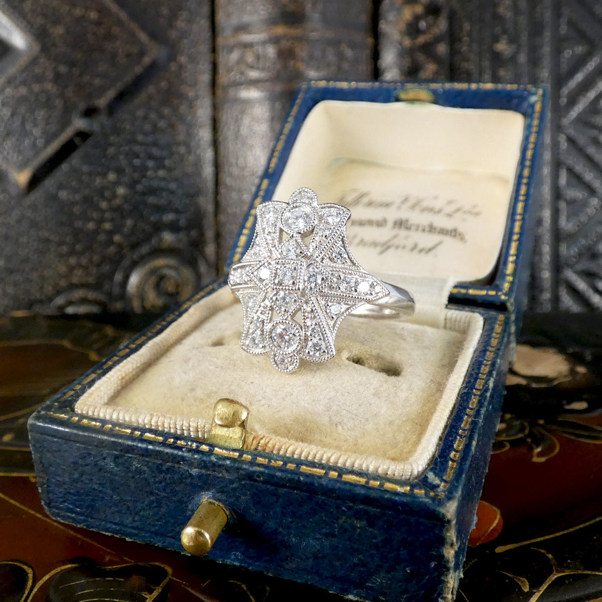 A platinum Art Deco style ring with pavé-set clear stones, displayed inside an open vintage blue velvet ring box with a cream cushion.