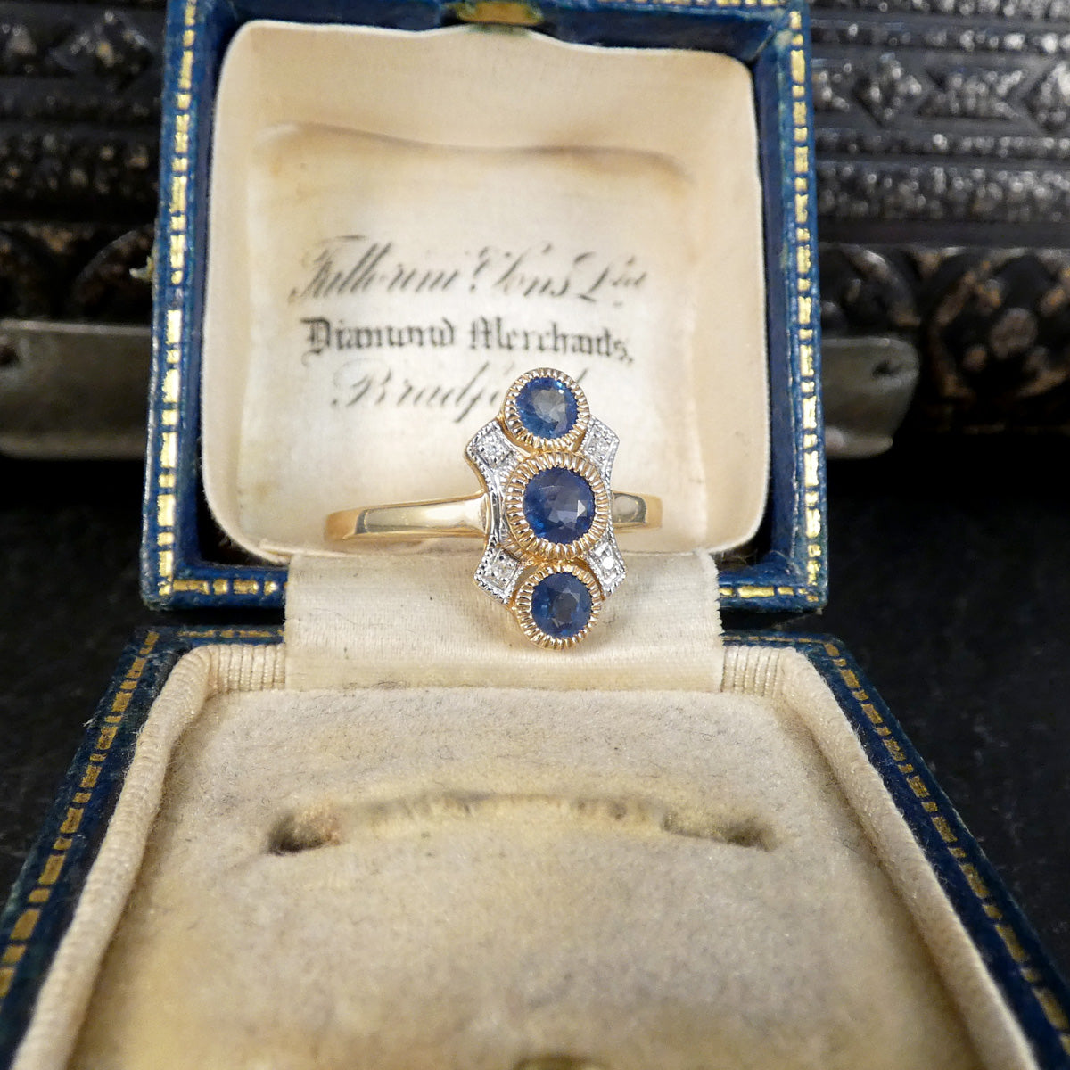 A vintage gold ring with three round blue sapphires and surrounding clear stones, displayed inside an open antique blue and gold jewelry box.