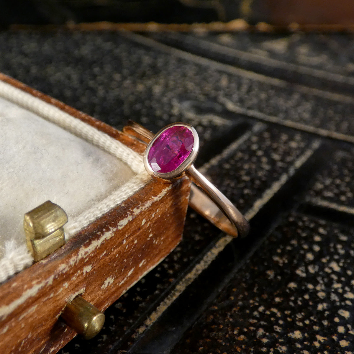 A rose gold ring with an oval-cut pink ruby gemstone resting on a vintage wooden jewelry box.