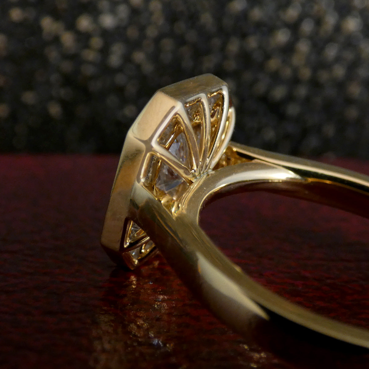 A close-up of a yellow gold ring featuring a geometric, fan-like setting with small clear stones, resting on a dark red textured surface with a dark, sparkly background.