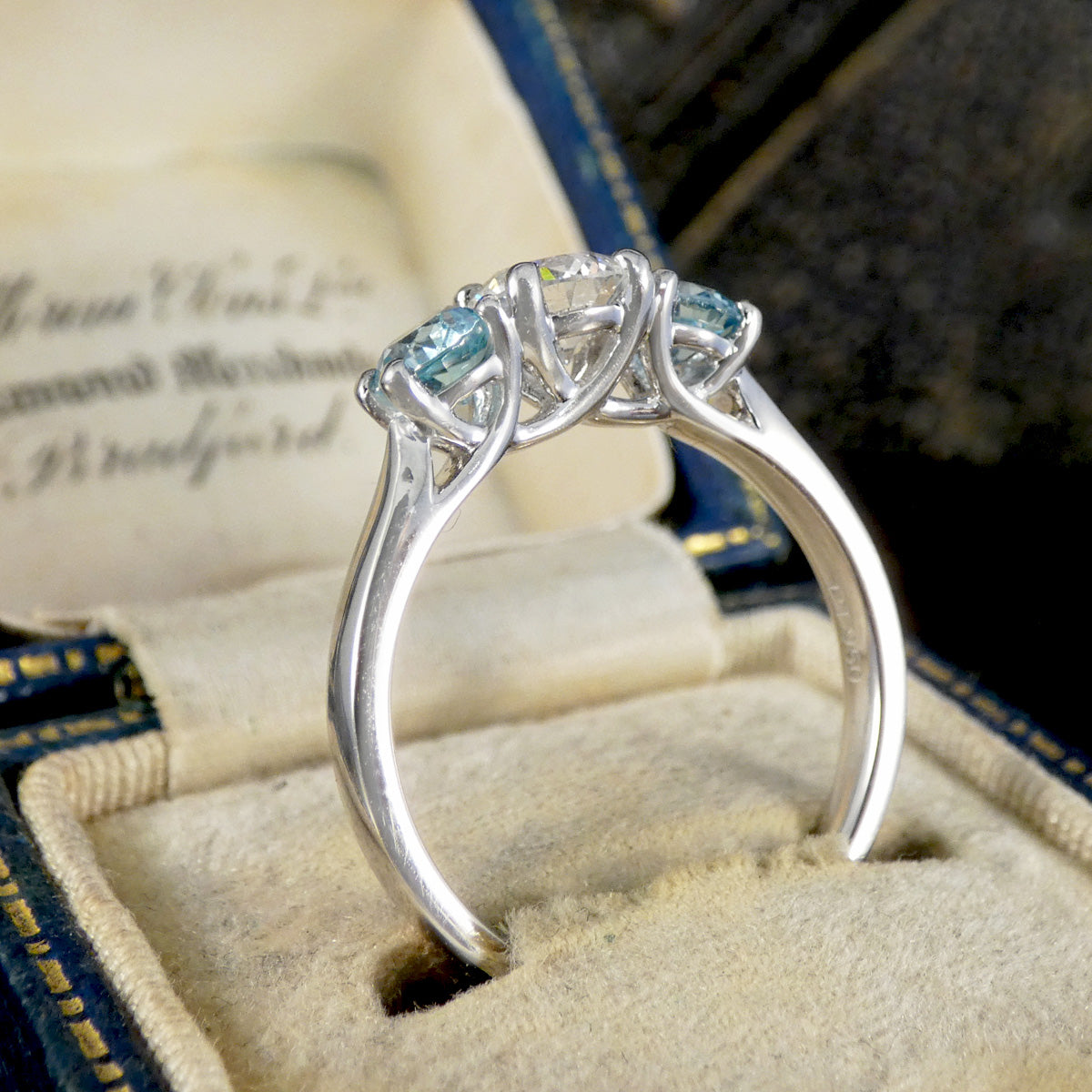 A silver three-stone ring with light blue gemstones displayed in a cream-colored velvet jewelry box.