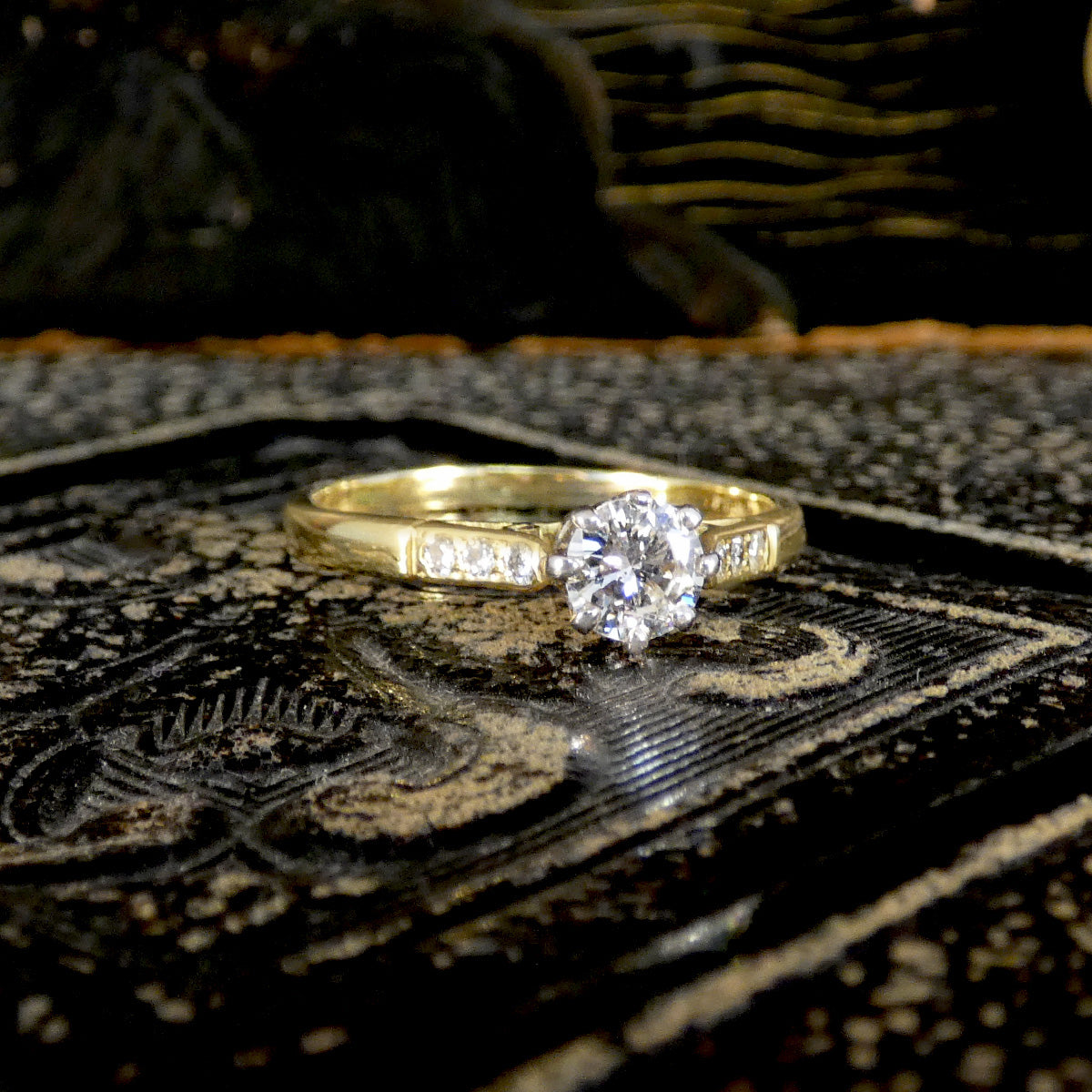 A yellow gold solitaire engagement ring with a round brilliant-cut diamond and small side diamonds, resting on a dark patterned book.